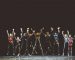 Group of young performers on the stage standing in the line, jumping with raised hands. Dark tones.
